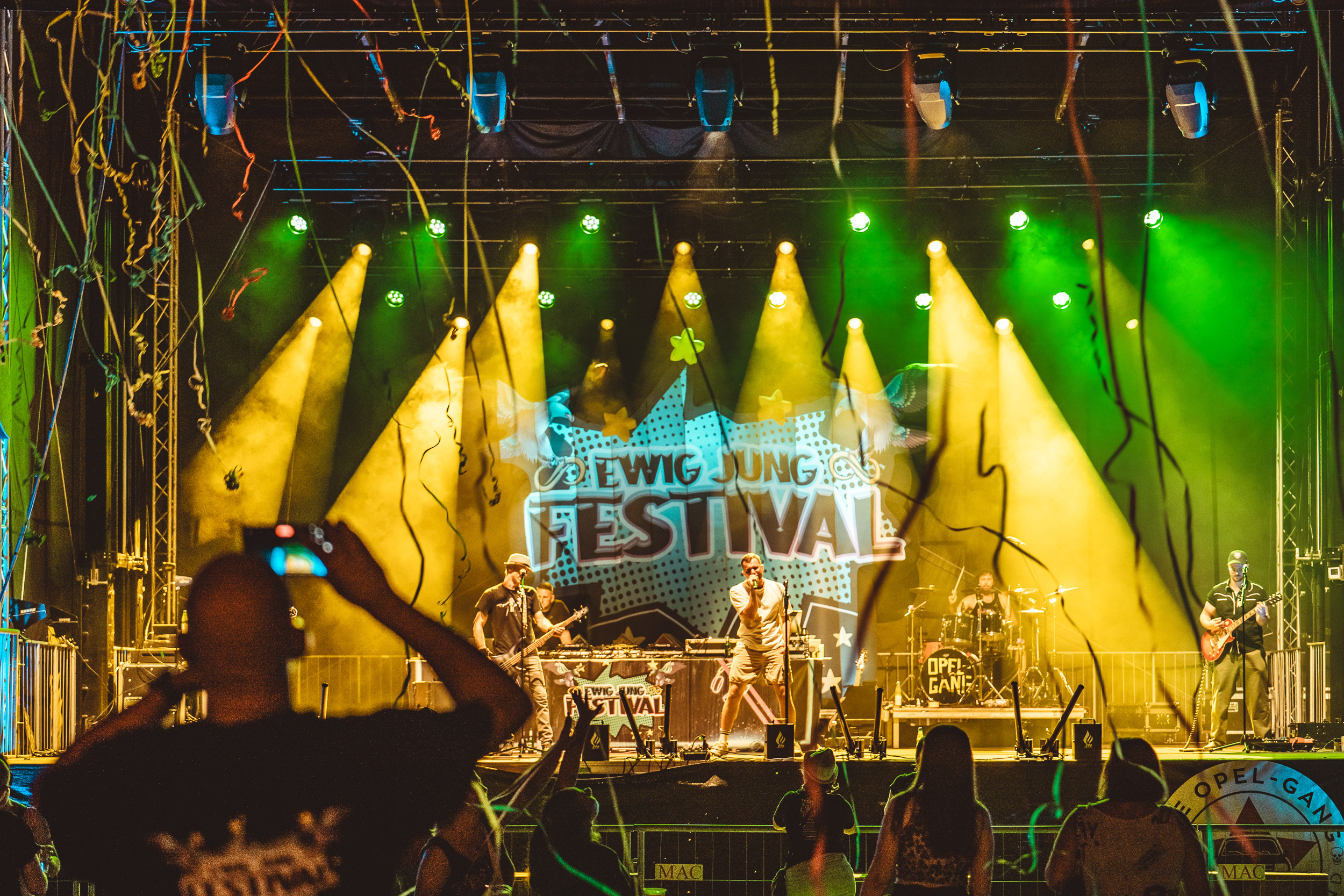 Ein Foto auf die Ewig Jung Festivalbühne in Thüringen frontal fotografiert. Man sieht i gelben Licht die Zuschauer und die komplette Band auf der Bühne.